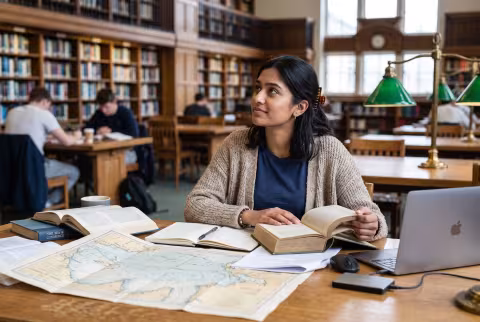 Research student at a library desk piled with charts and volumes — Person `main_image`, student.